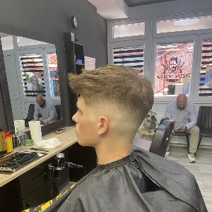 Klijent u frizerskom salonu za muškarce nakon šišanja. U pozadini vidljiv još jedan muškarac koji sjedi i čeka termin.