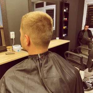 Na stolici nakon šišanja sjedi mlađi muškarac. U pozadini još dva muškarca čekaju svoj termin.